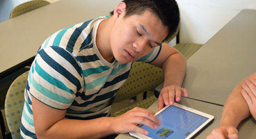 A young blind man leans forward as he listens to voiceover on an accessible iPad in a classroom.